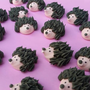 Puede incluir: Una colecci&oacute;n de dulces con forma de erizo con caras blancas, espaldas puntiagudas de color verde oscuro y peque&ntilde;os ojos y narices negros. Los dulces est&aacute;n dispuestos sobre una superficie de color p&uacute;rpura claro, creando una exhibici&oacute;n caprichosa y encantadora.