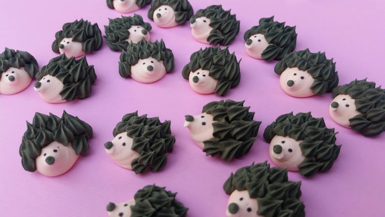 Puede incluir: Una colecci&oacute;n de dulces con forma de erizo con caras blancas, espaldas puntiagudas de color verde oscuro y peque&ntilde;os ojos y narices negros. Los dulces est&aacute;n dispuestos sobre una superficie de color p&uacute;rpura claro, creando una exhibici&oacute;n caprichosa y encantadora.