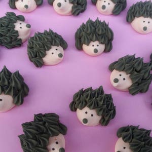 Puede incluir: Una colecci&oacute;n de golosinas con forma de erizo, con espinas de glaseado verde oscuro y caras blancas con ojos y narices de puntos negros. Las golosinas est&aacute;n dispuestas sobre una superficie de color lila claro.