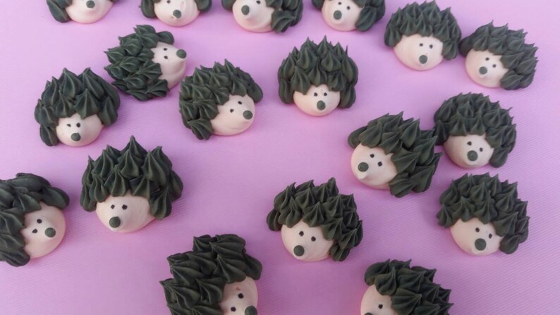 Puede incluir: Una colecci&oacute;n de golosinas con forma de erizo, con espinas de glaseado verde oscuro y caras blancas con ojos y narices de puntos negros. Las golosinas est&aacute;n dispuestas sobre una superficie de color lila claro.