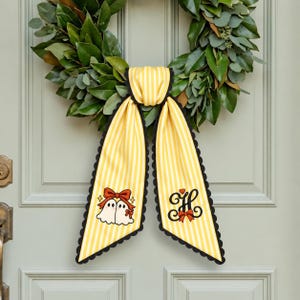 May include: A yellow and white striped fabric bow with black scalloped edges. One tail features a graphic of two white ghosts with red bows, and the other tail displays a black cursive letter H with a small red bow and heart accent.