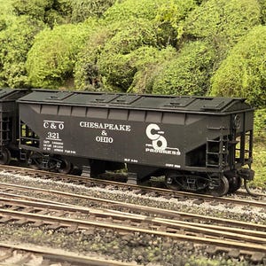 May include: A model train set featuring two black hopper cars with white lettering reading Chesapeake & Ohio and C&O Progress. The cars are positioned on metal tracks. Each car displays specific identification numbers 314 and 321 in white text.