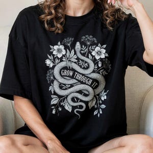 Könnte beinhalten: Ein schwarzes Kurzarm-T-Shirt mit einer weißen grafischen Illustration einer gewundenen Schlange, umgeben von botanischen Blumen und Blättern. Der Text "GROW THROUGH IT" ist in einer fetten, geschwungenen Schrift auf dem Schlangenkörper.