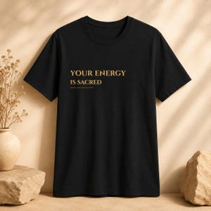 May include: A black short-sleeved t-shirt featuring the gold text "YOUR ENERGY IS SACRED" printed on the chest, with the smaller text "GOLD AURA COLLECTIVE" positioned directly underneath the main phrase in a clean, sans-serif font style.