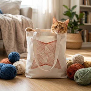 May include: A cream-colored canvas tote bag featuring a geometric red line art design on the front, with an orange tabby cat peeking out from the top opening, surrounded by several balls of yarn in various colors including blue, orange, and green.