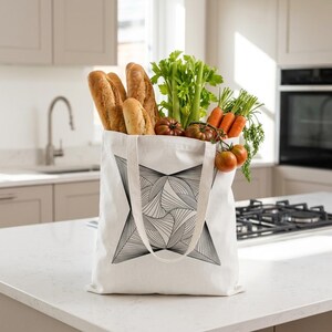 May include: A white canvas tote bag featuring a black geometric star-shaped line art design. The bag is filled with fresh groceries, including several long crusty bread loaves, a bunch of green celery, orange carrots, and red tomatoes.