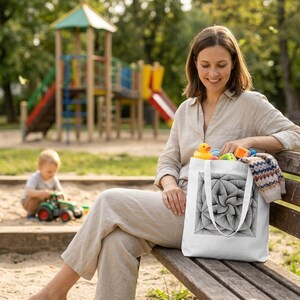 May include: A white canvas tote bag featuring a black geometric line art design of a swirling square pattern. The bag has two white fabric handles and is filled with various colorful plastic toys and a folded patterned knit garment.