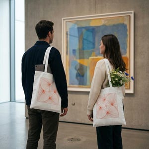 May include: Two white canvas tote bags featuring a square graphic print of orange radiating line patterns. The bags have two shoulder straps and are made of a light-colored fabric. One bag contains a bouquet of white and blue wildflowers.