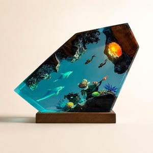 May include: A trapezoidal resin sculpture featuring an underwater scene with two dolphins, a sea turtle, and two scuba divers. The piece includes dark wood elements, coral-like textures, and a glowing orange light source, all mounted on a wooden base.