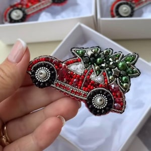 Può includere: Una spilla di perline a forma di auto rossa che trasporta un albero di Natale verde sul tetto. L'auto è decorata con perline rosse e nere, dettagli in filo argentato e ruote di perline bianche. L'albero presenta perline verdi e stelline.