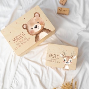 May include: Two rectangular wooden keepsake boxes featuring printed illustrations of a bear and a deer. The larger box displays the name MATHEO with birth details, while the smaller box shows the name AMELIE with birth details. Small wooden blocks are nearby.