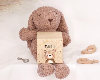 May include: A brown plush bunny toy holding a light-colored wooden cube. The cube features a printed illustration of a bear, small dots, a yellow banner, and the name MATTEO in black text. A small metal key and two wooden whale-shaped items are nearby.