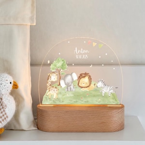 Może przedstawiać: Półkolista lampka nocna z akrylu z drewnianą podstawą, przedstawiająca akwarelową ilustrację żyrafy, słonia, lwa i zebry na zielonym polu. Tekst "Anton" oraz "30.10.2025" jest nadrukowany w kolorze białym nad zwierzętami.