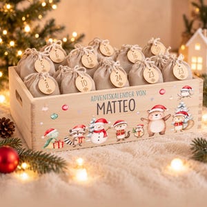 Peut inclure: Une caisse en bois rectangulaire contenant plusieurs petits sacs à cordon en tissu beige, chacun avec une étiquette en bois numérotée. L'avant de la caisse porte le texte "ADVENTSKALENDER VON MATTEO" et des illustrations d'animaux de Noël.