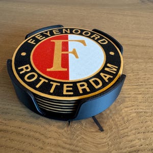 Op de afbeelding: Een stapel ronde onderzetters met het Feyenoord Rotterdam logo in rood, wit, zwart en goud, geplaatst in een zwarte kunststof houder. De bovenste onderzetter toont de letter F in goud op een gespleten rood-witte achtergrond.