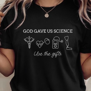 Op de afbeelding: Een zwart T-shirt met korte mouwen en de witte tekst GOD GAVE US SCIENCE en Use the gifts. Onder de tekst staan witte lijntekeningen van een caduceus, een hart met een ECG-lijn, een insulinepomp en een prothetisch been.