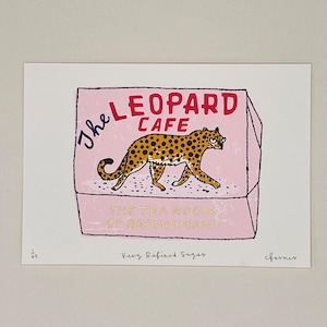 以下が含まれることがあります： 「The Leopard Cafe」の文字が赤と黒で描かれたピンク色の箱のプリント。箱の中には、黄色の毛皮と黒い斑点を持つヒョウがいます。「The Tea Room of Refinement」と「Very Refined Sugar」の文字も表示されています。