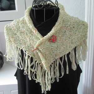 May include: A green and white knitted scarf with fringe on the edges. The scarf is draped over a black mannequin and has a wooden pin with a red and green button attached.