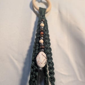 May include: A dark green macrame plant hanger featuring a light wood ring at the top. One strand is decorated with a vertical sequence of small round beads in silver, brown, and tan, ending with a large, oval, cream-colored stone bead with brown veining.