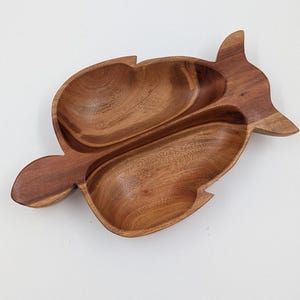 May include: A carved wooden serving dish shaped like a fish with two distinct, hollowed-out compartments separated by a central ridge. The wood features a rich, warm brown tone with visible natural grain patterns throughout the entire surface.