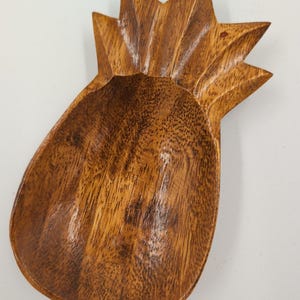 Puede incluir: Un cuenco de madera tallada con forma de piña y acabado liso y pulido. El cuenco presenta una base profunda y redondeada y una sección superior puntiaguda y texturizada que representa las hojas, mostrando un patrón de veta de madera natural.