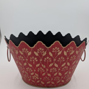 May include: A red metal bowl with a scalloped rim and a black interior. The exterior features a repeating gold floral pattern arranged in a diamond lattice design. Two small circular metal handles are attached to the sides of the bowl.