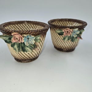 May include: Two ceramic planters featuring a woven basket texture in cream and brown. Each planter has a dark brown rope-style rim and a decorative relief of a pink rose, blue flowers, and green leaves attached to the front exterior surface.