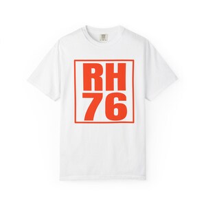 May include: A white short-sleeved t-shirt featuring a large, bold, orange square graphic on the chest. Inside the square, the text RH is printed on the top line and the number 76 is printed on the bottom line in a matching orange sans-serif font.
