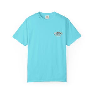May include: A bright turquoise short-sleeve t-shirt featuring a small circular graphic on the upper left chest. The graphic contains the text "BLESSED CREW" in a white and dark blue arched font, surrounded by small stars and decorative lines.