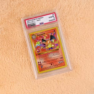 May include: A 1999 Pokemon French Dracaufeu Holo trading card graded GEM MT 10, encased in a clear protective plastic slab. The card features a Charizard illustration with a holographic background, red border, and printed game text in English.