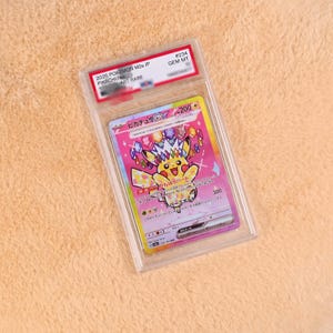 Könnte beinhalten: Eine bewertete 2025 Pokemon M2a JP Pikachu ex Special Art Rare Sammelkarte in einer durchsichtigen Kunststoffhülle. Die Karte zeigt ein gelbes Pikachu mit einer bunten, mit Edelsteinen besetzten Krone vor einem leuchtend rosa Hintergrund mit japanischem Text.