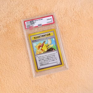 May include: A 1998 Japanese Promo Pikachu Illustrator trading card encased in a clear protective slab. The card features a yellow character holding a paintbrush, with the text ILLUSTRATOR and Japanese characters printed on a yellow and white background.