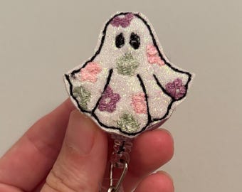 May include: A white glittery ghost-shaped badge reel featuring embroidered floral patterns in shades of pink, purple, and sage green. The ghost has two black embroidered eyes and a black stitched outline, attached to a clear plastic retractable cord.