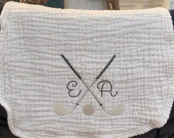 May include: A white muslin burp cloth featuring a textured, crinkled fabric surface. The centre is embroidered with two crossed golf clubs in dark grey thread, a small golf ball, and the cursive letters E and A in a matching dark grey tone.