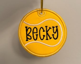 May include: A circular yellow embroidered ornament shaped like a tennis ball with white curved lines and the name Becky stitched in black thread across the centre. A small gold loop is attached at the top for hanging this decorative item.