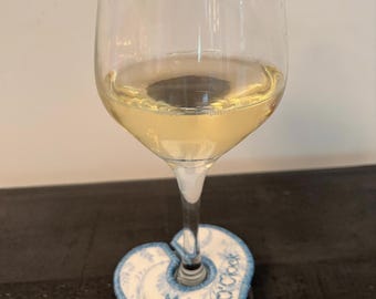 May include: A clear glass wine glass filled with a pale yellow liquid, featuring a heart-shaped fabric coaster at the base. The coaster is white with blue floral patterns and blue stitched edges, displaying the embroidered text Mom o'clock.