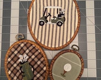 May include: Three oval embroidery hoops with wood-effect frames. One features a golf cart on striped fabric, another shows a golf bag on brown plaid fabric, and the third displays a golf ball on a tee against a solid light green fabric background.