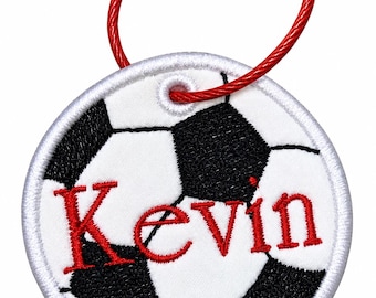May include: A circular embroidered patch featuring a black and white football pattern with the name Kevin stitched in red thread across the centre. The patch includes a white stitched border and a red metal cable loop attached at the top.