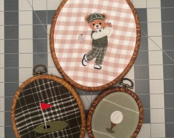 May include: Three oval embroidery hoops with faux wood frames. The largest features a teddy bear golfer on pink gingham fabric. The medium hoop shows a red flag on a green putting hole over dark plaid fabric. The smallest displays a golf ball on a tee.