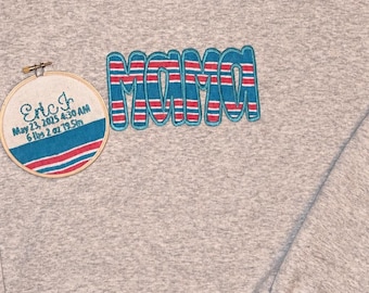 May include: A light grey heathered sweatshirt featuring the word MAMA embroidered in blue with red and white stripes. Beside it is a round wooden embroidery hoop displaying the text Eric Jr, May 23, 2025 4:30 AM, 6 lbs 2 oz 19.5 in.