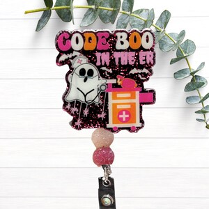 May include: A badge reel featuring a black glitter background with the text CODE BOO IN THE ER in pink and white. A white ghost wearing a nurse cap and glasses holds a stethoscope next to an orange medical cart with a pink cross symbol.
