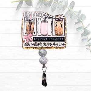 May include: A rectangular badge reel featuring a gold glitter border and a graphic illustration of three drinks. The text reads JUST A GIRL, STAYING HYDRATED, and with multiple drinks at a time. It includes two silver glitter beads and a black clip.