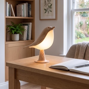 May include: A bird-shaped table lamp with a light wood grain finish on the head and base, featuring a glowing, translucent white body that emits a soft light. The lamp stands on a circular pedestal base and is positioned on a wooden desk surface.