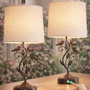 May include: A pair of table lamps with bronze-toned metal bases shaped like twisting branches featuring small birds and leaves. Each lamp has a cylindrical off-white fabric shade topped with a leaf-shaped finial and a circular base with a power button.