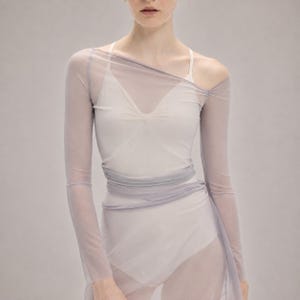 May include: A sheer, light lavender long-sleeve mesh top worn over a white camisole. The mesh fabric features an asymmetrical neckline that drapes across the chest and wraps around the waist, creating a layered effect with long, fitted sleeves.