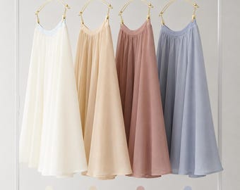 May include: Four pleated skirts hanging on gold-toned hangers on a white rack. The skirts are available in four colors: beige, cream, lotus pink, and slate blue. Each skirt features a flowing, lightweight fabric with a gathered waistband design.