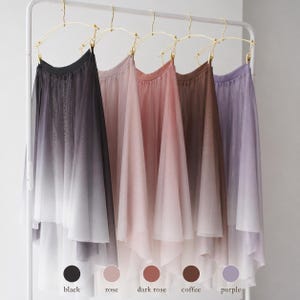 May include: Five midi-length skirts made of sheer, layered fabric hanging on gold-toned hangers. Each skirt features a vertical ombre gradient transitioning from a solid color at the waist to white at the hem. Colors include black, rose, dark rose, coffee, and purple.