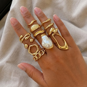 Op de afbeelding: Een verzameling goudkleurige ringen gedragen aan vier vingers. De ringen hebben diverse ontwerpen, waaronder gladde banden, getextureerde oppervlakken, kettingschakels en een grote, onregelmatige witte barokparel in een goudkleurige zetting.