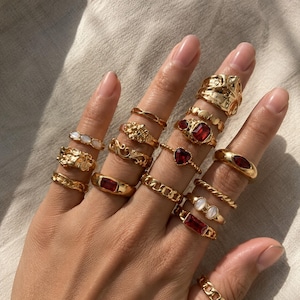 Può includere: Una collezione di anelli color oro indossati sulle dita, caratterizzati da vari design tra cui fasce testurizzate, fasce lisce, maglie a catena e anelli incastonati con pietre rosse rettangolari, a forma di cuore e ovali, oltre a piccoli accenti ovali bianchi.