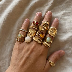 Può includere: Una collezione di anelli color oro indossati sulle dita, caratterizzati da vari design tra cui fasce lisce, superfici testurizzate e anelli con pietre rotonde o ovali nei toni del rosso, ambra, verde pallido e bianco, creando un aspetto stratificato.